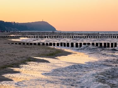 Strand auf Usedom mit Reihen von Holzbuhnen, die bei Sonnenuntergang in die Ostsee ragen. Im Hintergrund ist eine Klippe mit Leuchtturm sichtbar.