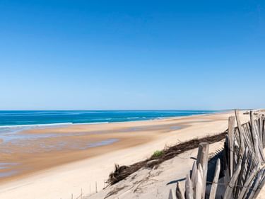 Breiter Sandstrand in Biscarosse mit Holzzaun auf Dünen im Vordergrund. Blauer Ozean und klarer Himmel erstrecken sich entlang der Küste.