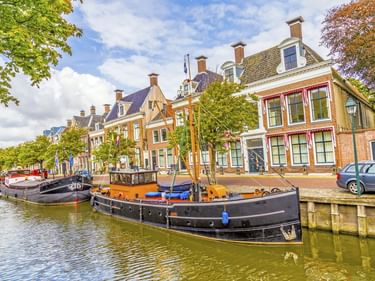 Traditionelle Holzboote liegen in einem Kanal in Harlingen, Niederlande, mit bunten historischen Gebäuden und Bäumen am Ufer.
