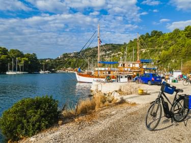 Fahrrad mit blauen Taschen auf Schotterweg neben ruhigem Hafen auf Paxos. Traditionelles Holzboot und Segelboote im Hafen mit grünen Hügeln.
