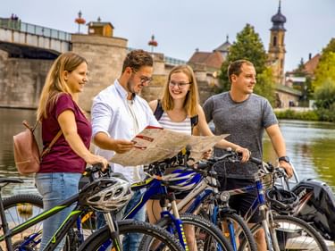 Four cyclists with bikes studying a map by the river in Kitzingen. Historic bridge and church tower visible in background.
