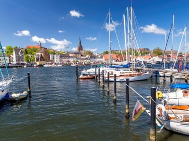 Flensburger Hafen mit vertäuten Segelbooten und Holzpfählen. Historische Gebäude und Kirchtürme sind über dem Wasser unter blauem Himmel sichtbar.