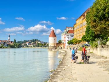 Cyclists on riverside path in Passau old town with Schaibling Tower and colorful historic buildings along the Inn River under blue sky.