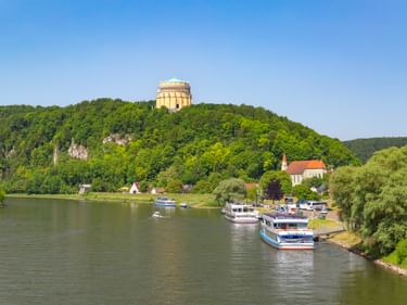 Befreiungshalle auf einem bewaldeten Hügel über der Donau in Kelheim, mit Ausflugsbooten am Ufer und einer sichtbaren Kirche.