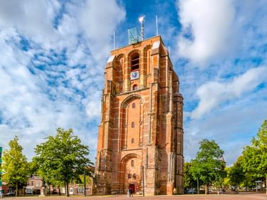 Der Oldehove, ein unvollendeter schiefer Backsteinturm in Leeuwarden, Niederlande, auf einem Platz umgeben von grünen Bäumen unter blauem Himmel.