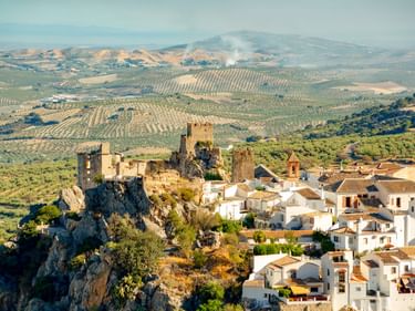 Zuheros Dorf mit weißen Häusern und mittelalterlicher Burgruine auf Felsvorsprung, umgeben von Hügeln mit Olivenhainen in Andalusien, Spanien.