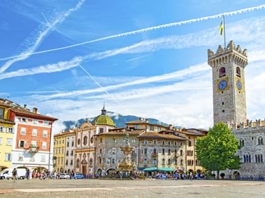 Piazza Duomo in Trient mit bunten historischen Gebäuden, Neptunbrunnen und Torre Civica Glockenturm unter blauem Himmel mit Kondensstreifen.