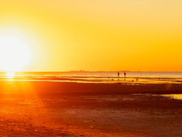 Goldener Sonnenuntergang über dem Strand von Cuxhaven mit Silhouetten von Menschen auf dem nassen Sand. Die Sonne erzeugt ein helles Leuchten am Horizont.