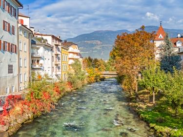 Fluss Rienz fließt durch Bruneck mit bunten Gebäuden auf beiden Seiten, Herbstbäumen und Bergen im Hintergrund unter blauem Himmel.