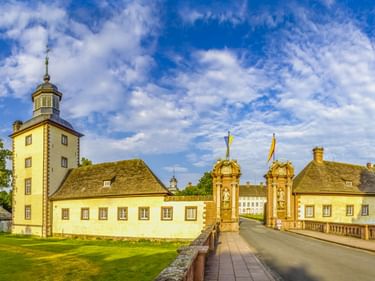 Schloss Corvey in Höxter with yellow tower building, ornate baroque gate pillars with flags, and white main palace visible beyond the bridge.