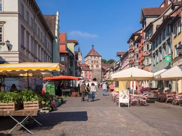 Fußgängerplatz in Rottweils Altstadt mit bunten historischen Gebäuden, Marktständen, Straßencafés und einem mittelalterlichen Turm im Hintergrund.