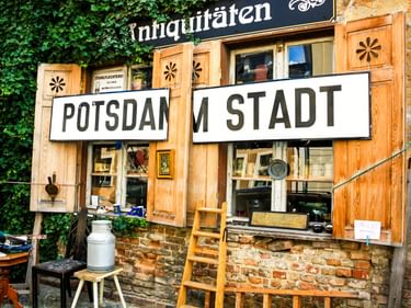 Rustikaler Antiquitätenladen mit Holzläden und Backsteinmauern. Große weiße Schilder mit 'POTSDAM STADT' über den Fenstern. Grüner Efeu oben.