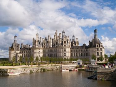 Schloss Chambord, ein Renaissance-Schloss mit markanten Türmen und Schornsteinen, vom Kanal aus gesehen mit Booten. Blauer Himmel mit weißen Wolken.