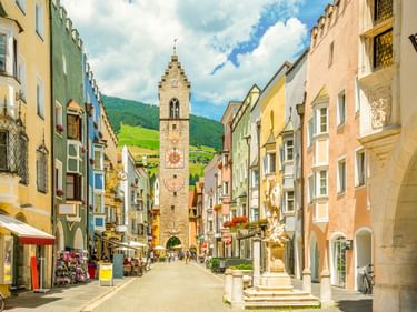 Bunte historische Gebäude säumen eine Fußgängerzone in Sterzing, Italien, mit einem hohen mittelalterlichen Turm in der Mitte und grünen Bergen im Hintergrund.