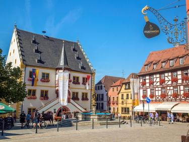 Volkacher Marktplatz mit historischem Rathaus, bunten Fachwerkhäusern und schmiedeeisernem Schild unter blauem Himmel.