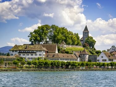 Blick auf Rapperswil vom Wasser mit weißen historischen Gebäuden am Ufer, einem Kirchturm auf einem Hügel umgeben von Bäumen und Bergen im Hintergrund unter blauem Himmel mit Wolken.
