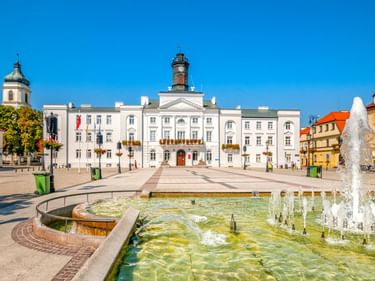 Weißes Rathaus mit Uhrturm in Plock, Polen. Großer Brunnen im Vordergrund auf sonnigem Platz mit bunten Gebäuden und Kirchturm.