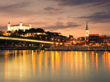 Bratislavaer Burg beleuchtet auf einem Hügel über der Donau bei Abenddämmerung. Stadtlichter spiegeln sich im Wasser, Martinsdom sichtbar.