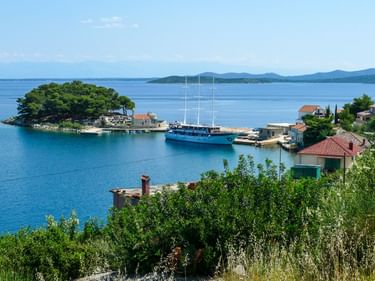 Küstenhafen auf Dugi Otok mit blauer Segelyacht am Pier, kleiner bewaldeter Insel, rotgedeckten Häusern und ruhiger blauer Adria.