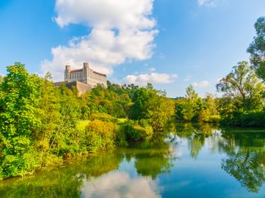 Willibaldsburg Castle on a hilltop in Eichstätt overlooking a calm river surrounded by lush green trees under a blue sky with white clouds.