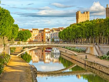 Kanal in Narbonne mit Steinbrücke, von Bäumen gesäumten Wegen und historischen Gebäuden. Mittelalterliche Türme im Hintergrund unter blauem Himmel.