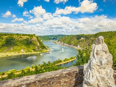 Stone statue of Loreley on rocky viewpoint overlooking the Rhine River bend with green forested hills, a cruise ship, and villages along the banks.