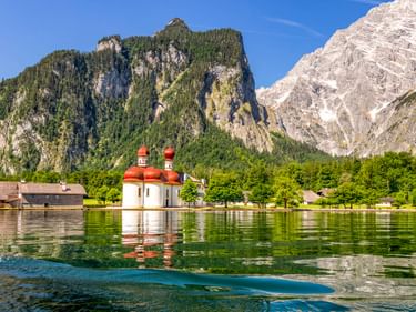 St. Bartholomä mit roten Zwiebeltürmen am Königssee, umgeben von Bergen. Klares Wasser spiegelt die Kirche und alpine Landschaft wider.