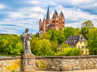 Steinstatue auf einer Brücke mit Blick auf den Limburger Dom mit seinen markanten roten Türmen über grünen Bäumen und einem gelben Gebäude an der Lahn.