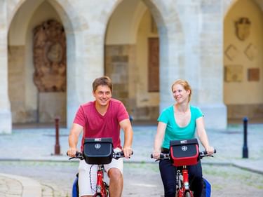 Zwei Radfahrer auf roten Rädern mit Lenkertaschen fahren auf Kopfsteinpflaster in Wien. Historisches Gebäude mit Torbögen im Hintergrund.