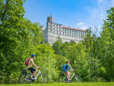 Zwei Radfahrer auf dem Altmühltal-Radweg durch grüne Wiese mit Willibaldsburg auf einem Hügel zwischen Bäumen unter blauem Himmel.