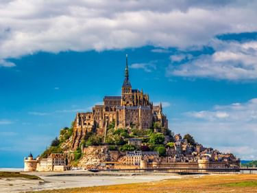 Mont Saint-Michel Abtei auf Felseninsel mit mittelalterlichen Gebäuden am Fuß, umgeben von Sandstrand und Wattenmeer unter bewölktem Himmel.