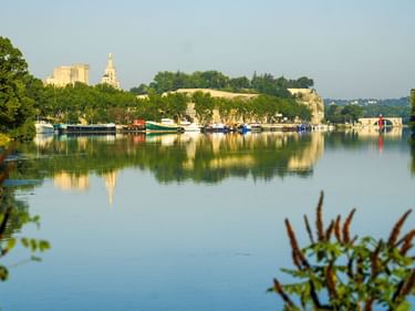 Ruhiger Flussblick in Avignon mit am Ufer vertäuten Booten. Historische Gebäude und Kirchturm auf dem Hügel, im Wasser gespiegelt.