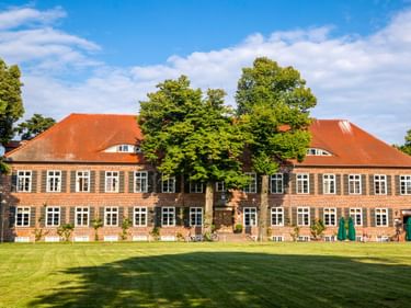 Gutshaus Ludorf, ein zweistöckiges Backsteingebäude mit rotem Ziegeldach und grünen Fensterläden, umgeben von hohen Bäumen und gepflegtem Rasen.