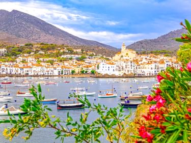 Hafen von Cadaqués mit bunten Fischerbooten, weißen Mittelmeerhäusern und Kirchturm. Rosa Blumen rahmen die Aussicht, Berge im Hintergrund.