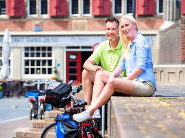 Zwei Radfahrer sitzen auf einer Steinmauer mit bepackten Tourenrädern auf einem Platz in einer Nordseestadt, historische Gebäude im Hintergrund.