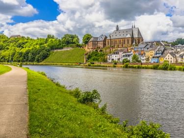 Radweg entlang der Saar in Saarburg mit historischen Gebäuden und Kirche am Hang. Weinberge und bunte Häuser unter bewölktem Himmel sichtbar.