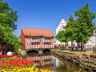 Orangefarbenes Fachwerkgebäude über einem Kanal im alten Hafen von Wismar. Bunte Blumen säumen das Ufer, dahinter ein weißer Kirchturm.