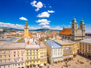 Aerial view of Linz old town with colorful buildings, baroque cathedral with twin towers, and yellow town hall tower under blue sky.