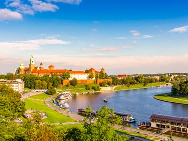 Luftaufnahme der Wawel-Burg mit rotem Dach auf einem Hügel an der Weichsel. Grüne Parkanlagen und Boote säumen das Flussufer in Krakau.