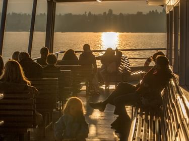 Passagiere auf dem Deck der Konstanz-Meersburg-Fähre bei Sonnenuntergang. Silhouetten von Menschen auf Bänken mit Bodensee und Ufer im Hintergrund.