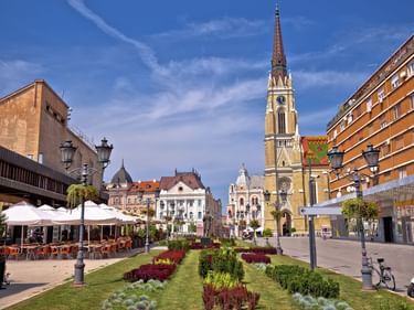 Sonniger Platz in Novi Sad mit hohem Kirchturm, bunten Gebäuden, Straßencafés und Grünanlagen unter blauem Himmel.