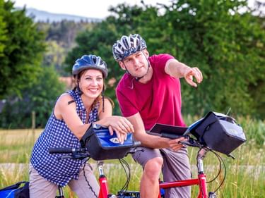 Two cyclists with helmets and panniers consulting a map on a rural path in the Dombes region, surrounded by green trees and fields.