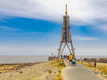 Die Kugelbake, ein hölzernes Seezeichen in Cuxhaven, mit Besuchern auf einem gepflasterten Weg. Sandstrand und Nordsee im Hintergrund.