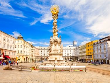 Weiße barocke Dreifaltigkeitssäule mit goldener Spitze am Hauptplatz in Linz, umgeben von bunten historischen Gebäuden unter blauem Himmel.
