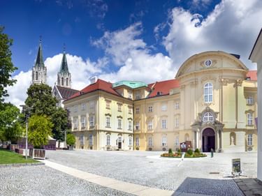 Stift Klosterneuburg mit gelben Barockgebäuden und zwei Kirchtürmen unter blauem Himmel. Gepflasterter Innenhof mit grünen Rasenflächen.