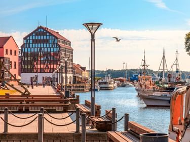 Hafenpromenade in Klaipeda mit historischen Fachwerkhäusern, Holzbooten und Schiffen am Kai unter blauem Himmel.