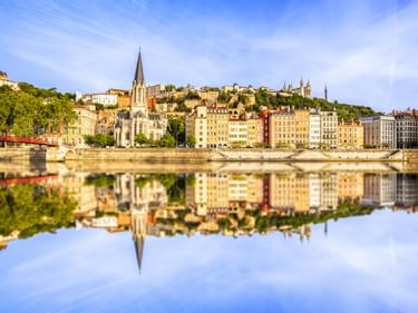 Kirche Saint-Georges mit hohem Turm in Lyons Altstadt spiegelt sich in der Saône. Bunte historische Gebäude säumen das Flussufer.