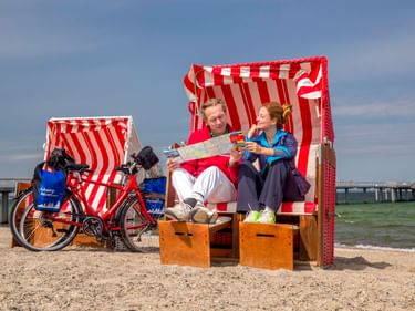 Zwei Radfahrer in einem rot-weiß gestreiften Strandkorb lesen eine Karte am Ostseestrand. Ein Tourenrad mit Taschen steht daneben.