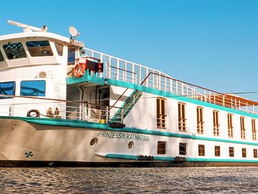 Flusskreuzfahrtschiff MS Prinzessin Katharina am Ufer. Weiß-türkisfarbenes Schiff mit mehreren Decks unter klarem blauen Himmel.
