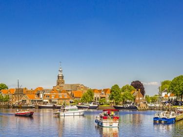 Malerischer Hafen von Blokzijl mit Booten auf ruhigem Wasser, historischen Gebäuden mit orangen Dächern und einem markanten Kirchturm unter blauem Himmel.